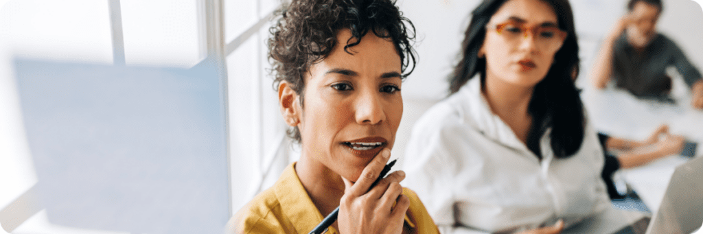 A professional woman in a yellow shirt holding a pen, deeply focused during a discussion, representing thoughtful decision-making in marketing strategy.