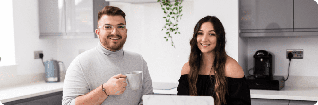 The Business Tea team, Ross and Sarah Powell, are holding mugs and smiling at the camera in a relaxed, modern work environment.