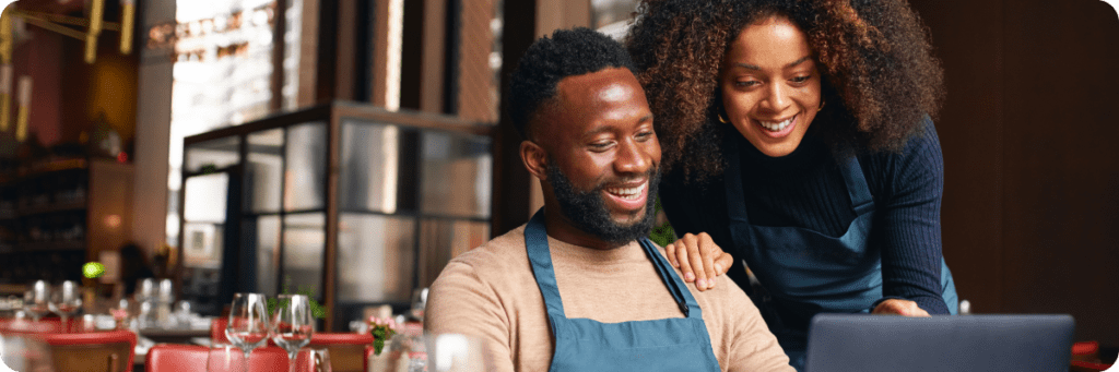 A smiling man and woman, both wearing aprons, looking at a laptop in a restaurant setting, representing small business owners using social media tools.