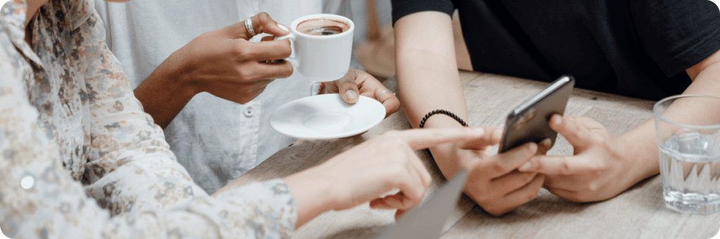 A group of three people sitting at a table, holding coffee cups, pointing at a smartphone, and discussing social media strategies for small businesses.